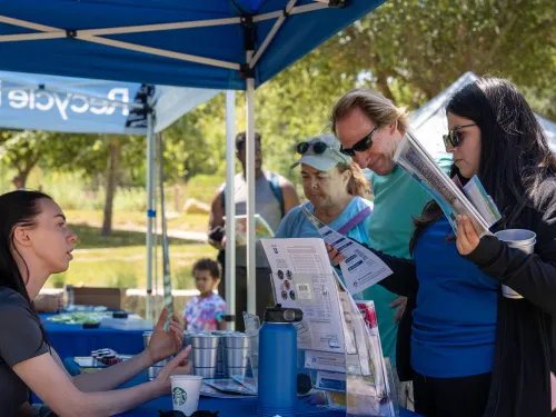 Vendor at a blue outdoor booth talks to two visitors holding pamphlets on a sunny day.