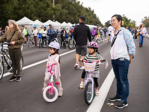 Two young girls in helmets on bikes with adults at an outdoor cycling event.