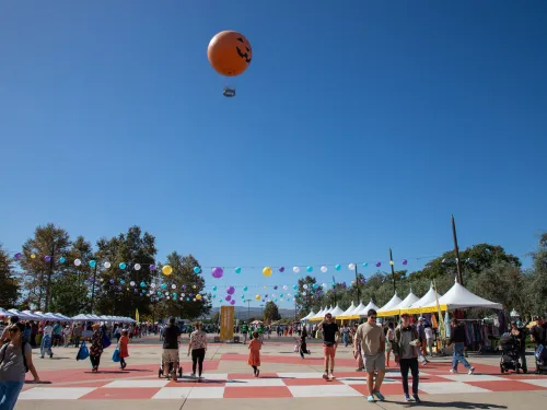 Outdoor festival with people, white vendor tents, and a smiling orange hot air balloon in a clear blue sky.