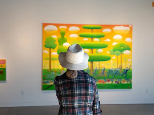 Person in a hat viewing a large, colorful painting of a stylized landscape in an art gallery.