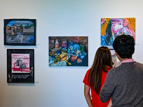 Two people from behind observe five colorful paintings in an art gallery.