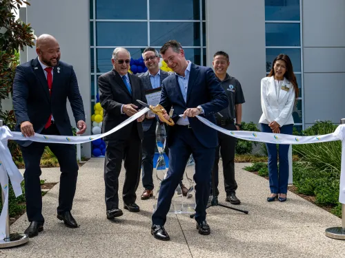 Man in suit cutting a ceremonial ribbon outdoors, surrounded by smiling people.