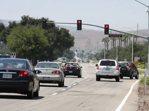 Cars wait at red traffic lights on a multi-lane road with trees and hills under a sunny sky.