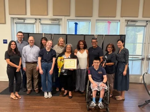 Photograph: Diverse group smiling, celebrating woman with award and flowers. Young man in wheelchair.