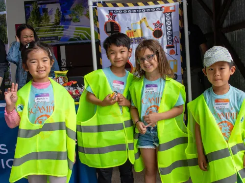 Four smiling children in bright yellow safety vests pose at a public works event.