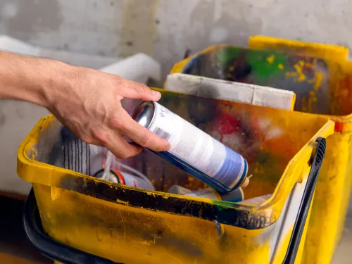 Hand places a spray paint can into a yellow, paint-splattered bucket.