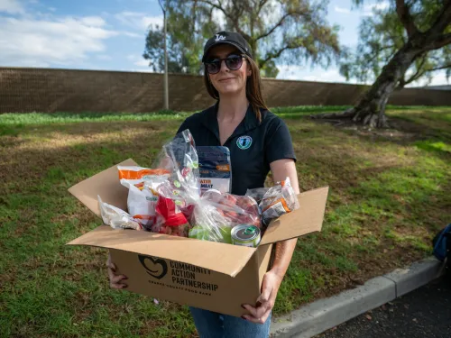 Smiling woman in a cap holds a full box of groceries outdoors.