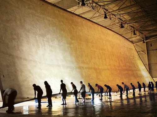 Photograph of workers collaboratively unrolling an enormous beige fabric in a vast hangar.