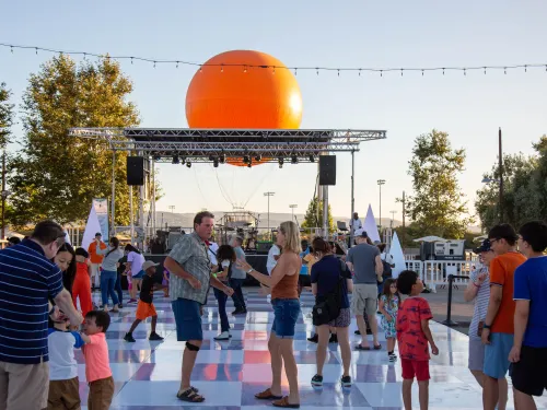 Outdoor event with people dancing on a checkered floor, large orange sphere behind stage.