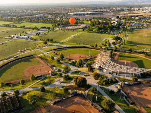 Aerial of a large park with baseball fields, green trees, and a distant orange hot air balloon.