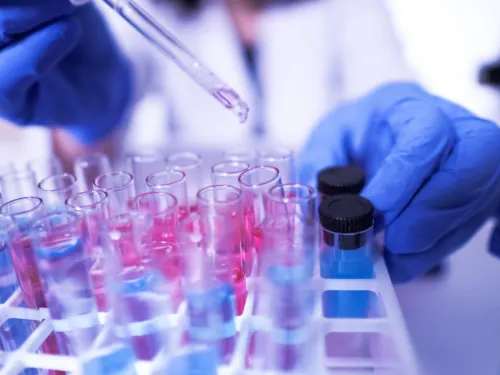 Scientist in blue gloves drips liquid from a pipette into a test tube among colorful samples.