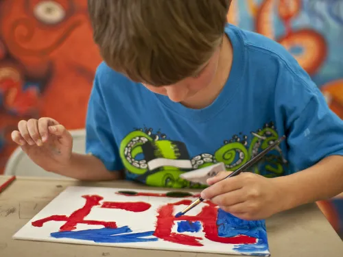 Boy focused on painting red and blue abstract shapes on paper, colorful mural behind him.