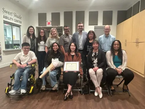 Diverse group of 12 people, two in wheelchairs, smiling with a certificate and flowers.
