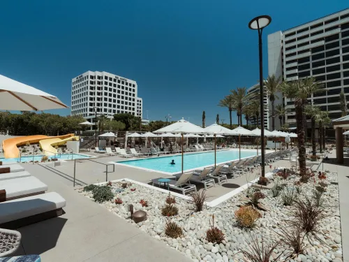 Large resort pool with lounge chairs, white umbrellas, a water slide, and tall buildings.