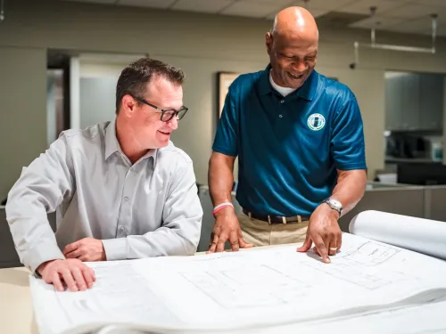 Photograph: Two smiling men review architectural blueprints in an office; one points.