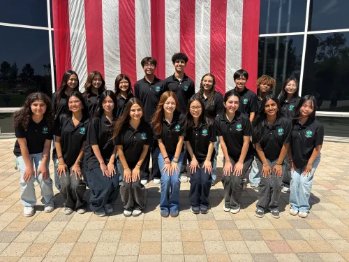 Smiling group of 20 young people in black shirts before a large American flag.